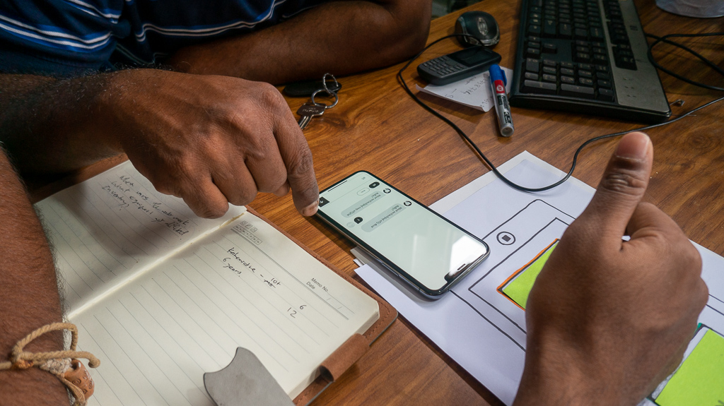 In-field user testing session: an SME participant at a desk pointing to the prototype on an Android phone beside a notebook and hand-drawn app wireframes, illustrating the co-creation workshop setup.