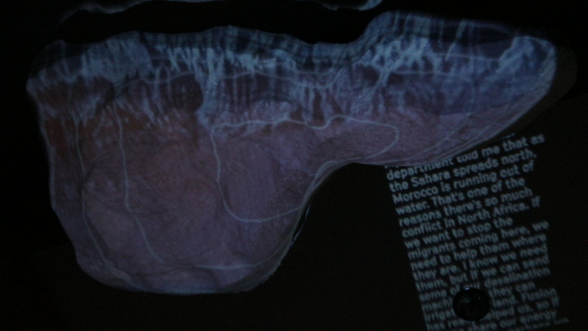 A topographic data-slice of North Africa is projected onto a curved surface, overlaid with scrolling policy narratives about water scarcity and migration—one of the three synchronized “mission slices” that anchor each loop’s storyline.